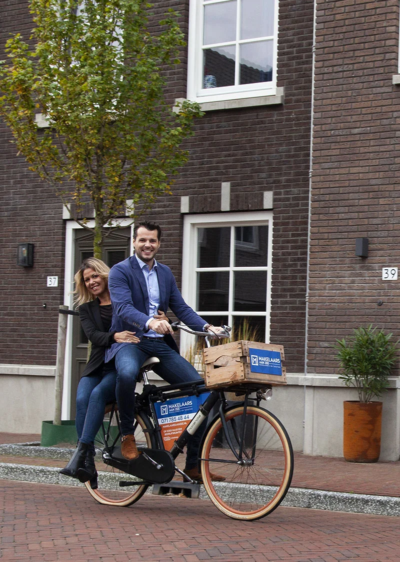 Makelaar uit Noordwijk fietsend door een straat met een vrouw achterop